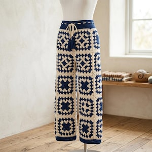 May include: Cream and navy blue crochet pants. The pants feature a geometric pattern of navy blue squares within cream-colored squares. The waistband is navy blue with a drawstring and tassel. The pants are displayed on a mannequin.