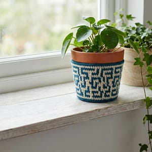 May include: A terracotta plant pot with a green plant, encased in a blue and cream crochet cozy. The cozy features a geometric pattern. The pot sits on a white windowsill, with a blurred background.