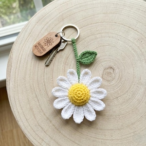 Op de afbeelding: Een handgemaakte sleutelhanger met een madeliefje van wit gehaakt garen, een gele kern en een groene stengel en blad. De sleutelhanger bevat een houten label met de tekst "HANDMADE WITH LOVE" en een zilveren sleutelring.