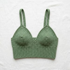 Può includere: Un top all'uncinetto verde salvia con scollo a V e spalline larghe. Il top fatto a mano presenta una fascia a coste sul fondo e un motivo a punto strutturato. Il top è presentato su sfondo bianco.