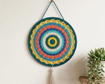 May include: A colorful, round crochet wall hanging with a mandala design. The piece features concentric circles in shades of teal, yellow, orange, blue, and cream. A cream-colored tassel hangs from the bottom. The wall hanging is suspended by a teal chain.
