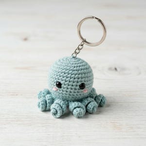 May include: A crocheted octopus keychain in a soft teal color. The octopus has a round head, eight curled tentacles, and black eyes with pink blush. A silver keyring is attached to the top of the octopus's head.