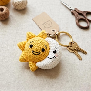 Pode incluir: Um chaveiro de crochê feito à mão com um sol amarelo sorridente e uma lua branca sorridente. O chaveiro é preso a um anel dourado com duas chaves e uma etiqueta que diz "HANDMADE SUN & MOON".