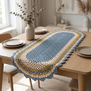 May include: A handmade, oval-shaped crochet table runner. The runner features a blue, cream, and yellow color scheme with a scalloped edge. The open weave design is visible, and the runner is placed on a wooden table.
