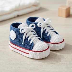 May include: A pair of handmade crochet baby trainers in navy blue and white. These high-top style shoes feature white laces, a white toe cap, and a thin red stripe encircling the white sole. A circular white patch is visible on the side.