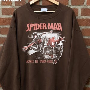 Puede incluir: Sudadera marr&oacute;n con un gr&aacute;fico de Spider-Man. El texto "SPIDER-MAN" es rosa y "ACROSS THE SPIDER-VERSE" es blanco. Tiene cuello redondo y mangas largas. La palabra "SWEATSHIRT" est&aacute; en la parte superior.
