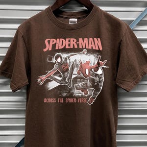 Puede incluir: Camiseta marr&oacute;n chocolate oscuro con el logo de Spider-Man en rosa y blanco, con dos personajes de Spider-Man en acci&oacute;n. El texto "ACROSS THE SPIDER-VERSE" est&aacute; debajo. La camiseta est&aacute; colgada en una percha de madera.