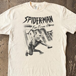 K&ouml;nnte beinhalten: Cremefarbenes T-Shirt mit einer Grafik von Spider-Man in dynamischer Pose und dem Text "SPIDERMAN Far From Home" in Schwarz. Das Design beinhaltet eine Graustufen-Illustration einer Stadtlandschaft. Das Shirt hat kurze &Auml;rmel und einen Rundhalsausschnitt.
