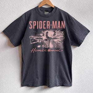 May include: Dark gray t-shirt featuring the words "SPIDER-MAN" in pink above a graphic of the superhero with outstretched arms. The word "Homecoming" is printed in pink script below the graphic. The shirt is hanging on a wooden hanger.