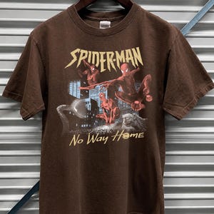 Może przedstawiać: Ciemnobrązowy t-shirt w kolorze gorzkiej czekolady z grafiką Spider-Mana. Projekt zawiera napis "SPIDER-MAN" złotymi literami oraz frazę "No Way Home". Grafika przedstawia Spider-Mana w akcji na tle miejskiego krajobrazu.