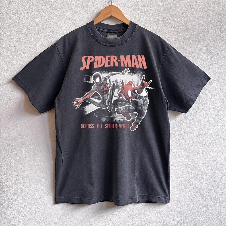 Puede incluir: Camiseta gris oscuro con un gr&aacute;fico de Spider-Man. El dise&ntilde;o incluye las palabras "SPIDER-MAN" en rosa sobre una ilustraci&oacute;n de dos personajes de Spider-Man. El texto "ACROSS THE SPIDER-VERSE" est&aacute; impreso debajo.
