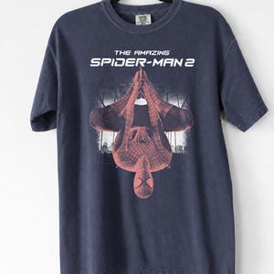 May include: Navy blue t-shirt featuring the text "THE AMAZING SPIDER-MAN 2" above a red and black image of Spider-Man. The superhero is depicted upside down with a cityscape in the background. The shirt is hanging on a black hanger.