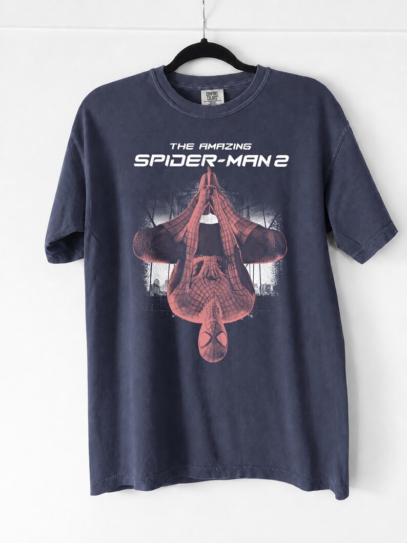 May include: Navy blue t-shirt featuring the text "THE AMAZING SPIDER-MAN 2" above a red and black image of Spider-Man. The superhero is depicted upside down with a cityscape in the background. The shirt is hanging on a black hanger.