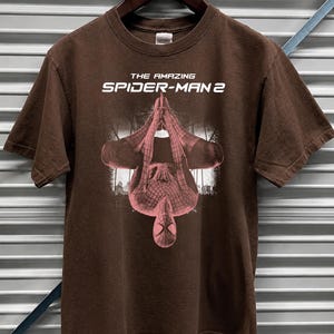May include: Dark chocolate brown t-shirt featuring the text "THE AMAZING SPIDER-MAN 2" in white. The shirt displays a pink and red graphic of Spider-Man. The shirt is hanging on a wooden hanger.