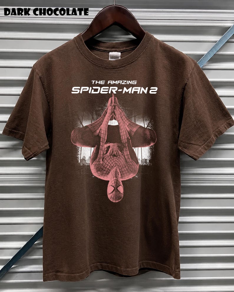 May include: Dark chocolate brown t-shirt featuring the text "THE AMAZING SPIDER-MAN 2" in white. The shirt displays a pink and red graphic of Spider-Man. The shirt is hanging on a wooden hanger.