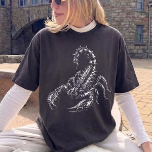 May include: A charcoal gray short-sleeve t-shirt featuring a large, detailed white graphic print of a scorpion on the front. The shirt has a crew neckline and is layered over a long-sleeve white shirt with a high neck, creating a contrasting look.