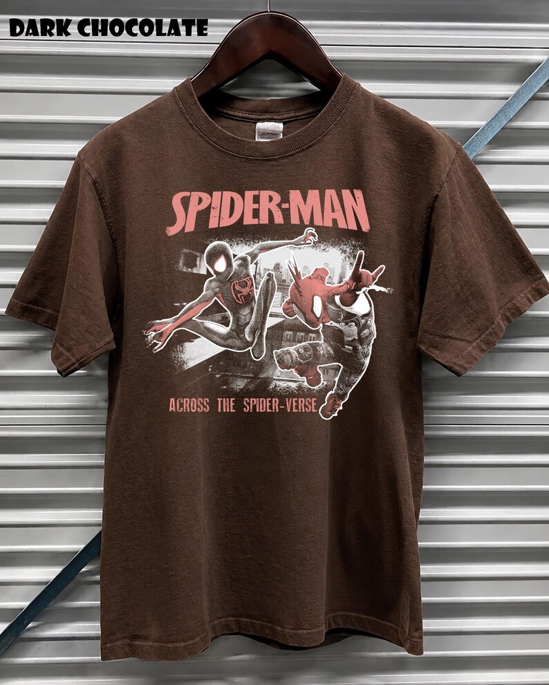 Puede incluir: Camiseta marr&oacute;n chocolate oscuro con el logo de Spider-Man en rosa y blanco, con dos personajes de Spider-Man en acci&oacute;n. El texto "ACROSS THE SPIDER-VERSE" est&aacute; debajo. La camiseta est&aacute; colgada en una percha de madera.