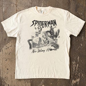 Może przedstawiać: Kremowa koszulka z czarno-białą grafiką Spider-Mana w akcji, z napisem "SPIDER-MAN" nad obrazem i "No Way Home" poniżej. Koszulka wykonana jest z miękkiego materiału.