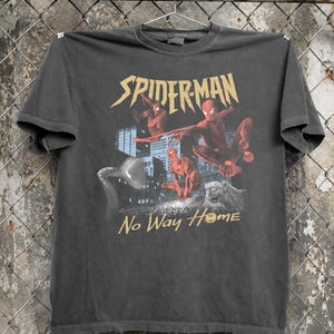Może przedstawiać: Ciemnoszara koszulka z grafiką Spider-Mana i złotym napisem "SPIDERMAN" nad obrazem. Słowa "No Way Home" są wydrukowane na złoto poniżej. Koszulka wisi na metalowym wieszaku.
