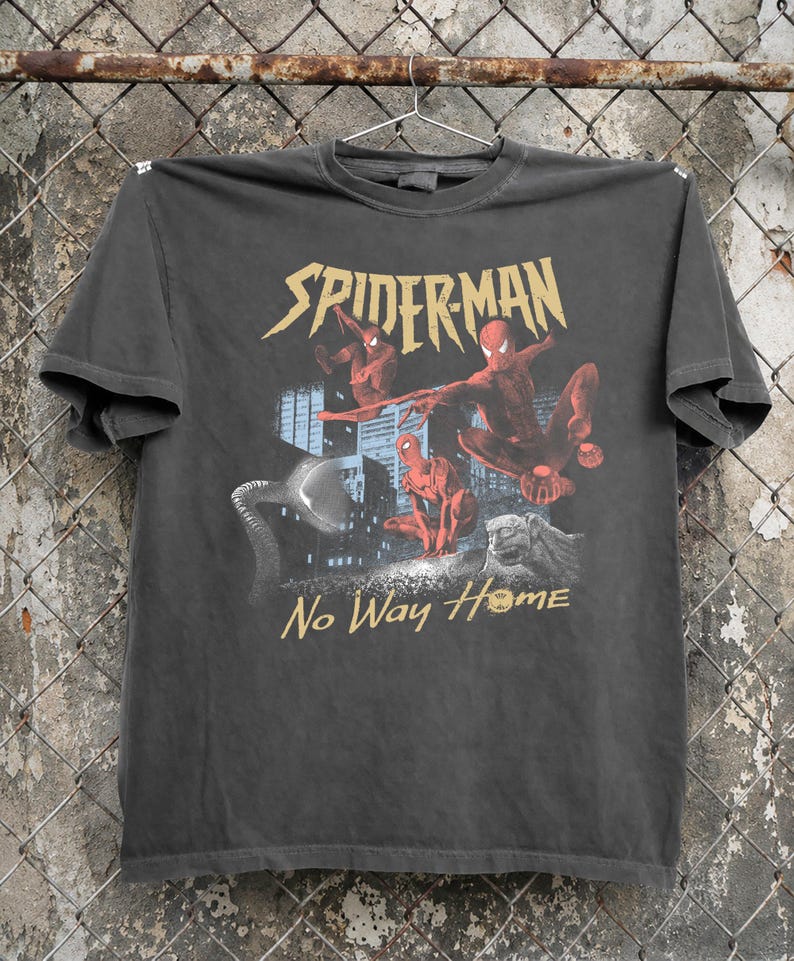 Może przedstawiać: Ciemnoszara koszulka z grafiką Spider-Mana i złotym napisem "SPIDERMAN" nad obrazem. Słowa "No Way Home" są wydrukowane na złoto poniżej. Koszulka wisi na metalowym wieszaku.