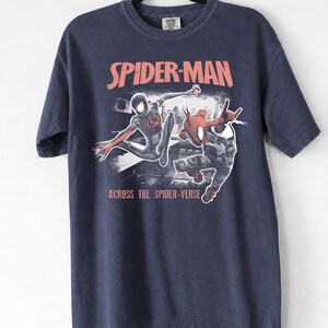 Puede incluir: Camiseta azul marino con un dise&ntilde;o naranja de Spider-Man y el texto "SPIDER-MAN" encima de la imagen y "ACROSS THE SPIDER-VERSE" debajo. La camiseta est&aacute; colgada de una percha negra.
