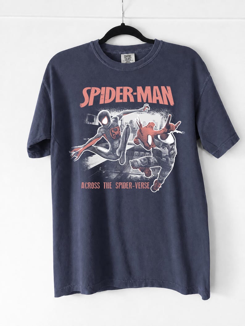 Puede incluir: Camiseta azul marino con un dise&ntilde;o naranja de Spider-Man y el texto "SPIDER-MAN" encima de la imagen y "ACROSS THE SPIDER-VERSE" debajo. La camiseta est&aacute; colgada de una percha negra.