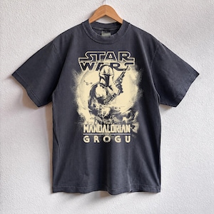 May include: Dark gray t-shirt featuring a graphic from the Star Wars series, with the text "STAR WARS" above an image of The Mandalorian holding a weapon. The text "THE MANDALORIAN AND GROGU" is below the image.