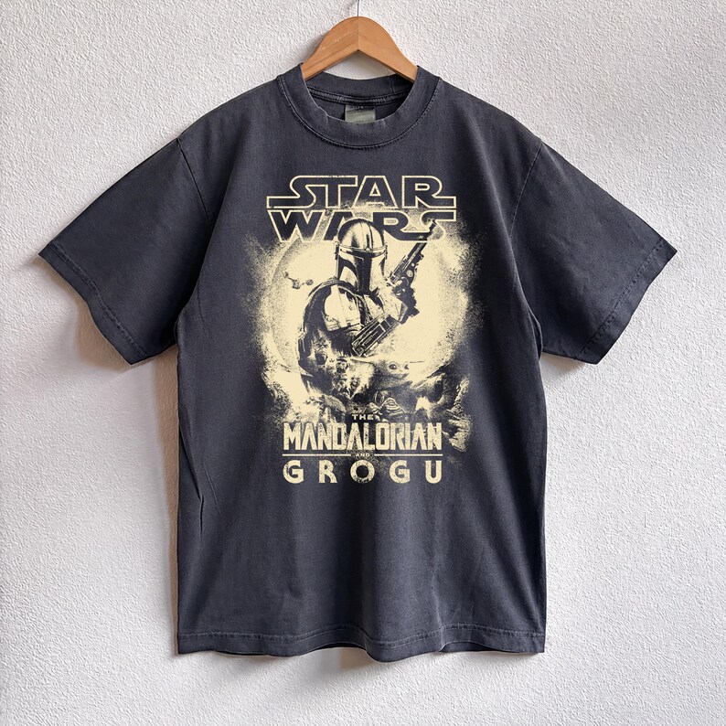 May include: Dark gray t-shirt featuring a graphic from the Star Wars series, with the text "STAR WARS" above an image of The Mandalorian holding a weapon. The text "THE MANDALORIAN AND GROGU" is below the image.