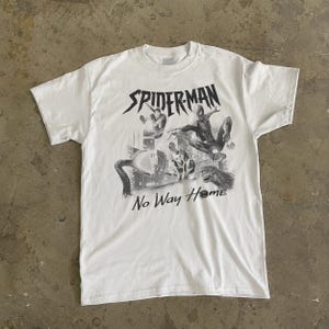 Może przedstawiać: Biała koszulka z czarno-białą grafiką postaci Spider-Mana i napisem "SPIDERMAN No Way Home". Projekt zawiera wiele postaci Spider-Mana w pozach akcji na tle miejskiego krajobrazu.