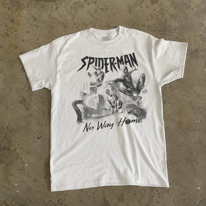 Może przedstawiać: Biała koszulka z czarno-białą grafiką postaci Spider-Mana i napisem "SPIDERMAN No Way Home". Projekt zawiera wiele postaci Spider-Mana w pozach akcji na tle miejskiego krajobrazu.