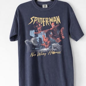 Może przedstawiać: Granatowa koszulka z nadrukiem Spider-Mana w akcji, złotymi napisami "SPIDERMAN" i "No Way Home", budynkami miejskimi i kamiennym gargulcem.