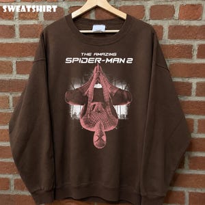 May include: Brown sweatshirt featuring a graphic of Spider-Man from "The Amazing Spider-Man 2" movie. The design shows Spider-Man upside down with a cityscape background. The word "SWEATSHIRT" is at the top.