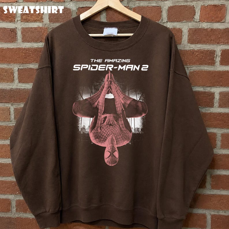 May include: Brown sweatshirt featuring a graphic of Spider-Man from "The Amazing Spider-Man 2" movie. The design shows Spider-Man upside down with a cityscape background. The word "SWEATSHIRT" is at the top.