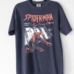 K&ouml;nnte beinhalten: Marineblaues T-Shirt mit einem Spider-Man-Motiv aus dem Film "Far From Home." Die Grafik zeigt Spider-Man in einer dynamischen Pose vor einer Stadtkulisse. Der Text "SPIDER-MAN Far From Home" ist rot gedruckt.
