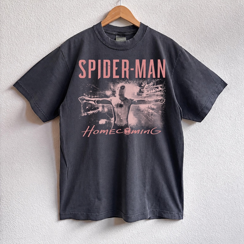May include: Dark gray t-shirt featuring the words "SPIDER-MAN" in pink above a graphic of the superhero with outstretched arms. The word "Homecoming" is printed in pink script below the graphic. The shirt is hanging on a wooden hanger.
