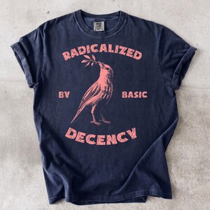 Puede incluir: Una camiseta de manga corta en color gris carbón oscuro con un gráfico rosa desgastado de un pájaro que sostiene una pequeña rama con hojas en el pico. El texto RADICALIZED BY BASIC DECENCY está impreso en rosa alrededor del ave.