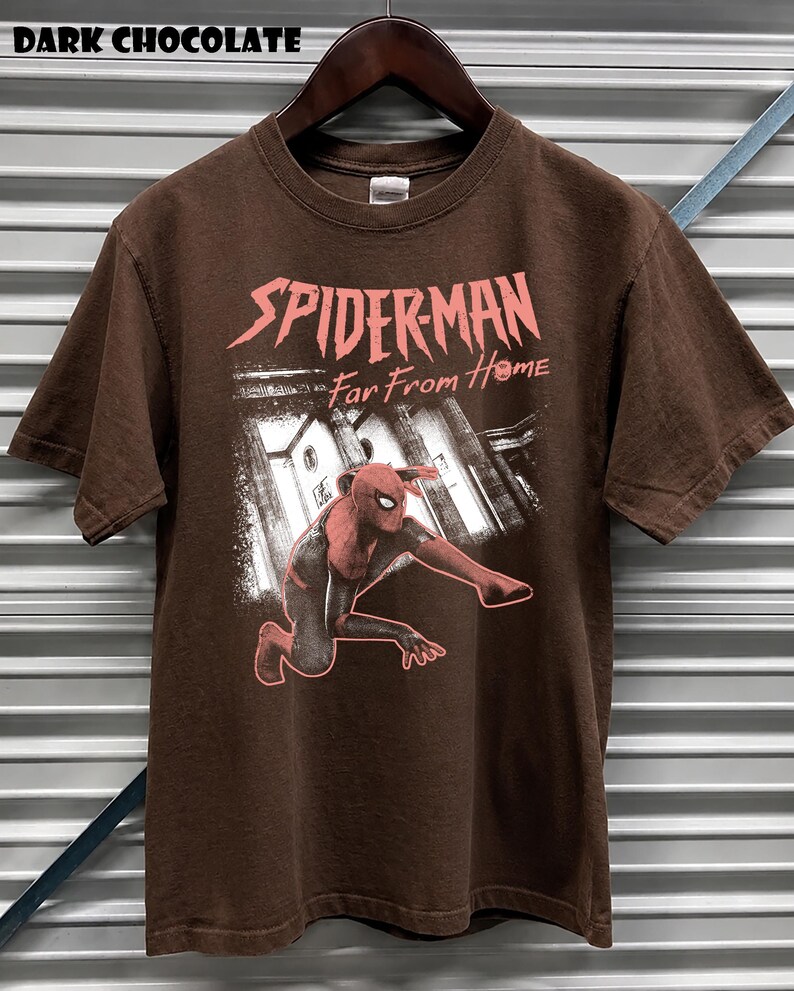 K&ouml;nnte beinhalten: Dunkelbraunes T-Shirt mit dem Text "SPIDERMAN Far From Home" in Rosa. Das Shirt zeigt eine Illustration von Spider-Man in einer dynamischen Pose. Das T-Shirt h&auml;ngt an einem Holzb&uuml;gel.