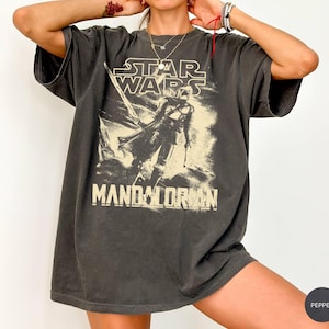May include: Dark gray Star Wars Mandalorian graphic t-shirt. The shirt features the Star Wars logo and a graphic of the Mandalorian. The shirt is oversized with short sleeves.