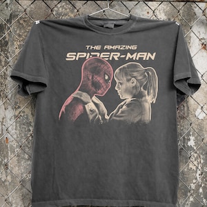 K&ouml;nnte beinhalten: Dunkelgraues T-Shirt mit einer Grafik von Spider-Man und einer Frau, die sich gegen&uuml;berstehen. Der Text "THE AMAZING SPIDER-MAN" ist in hellem Beige &uuml;ber dem Bild aufgedruckt. Das T-Shirt h&auml;ngt an einem Metallb&uuml;gel.