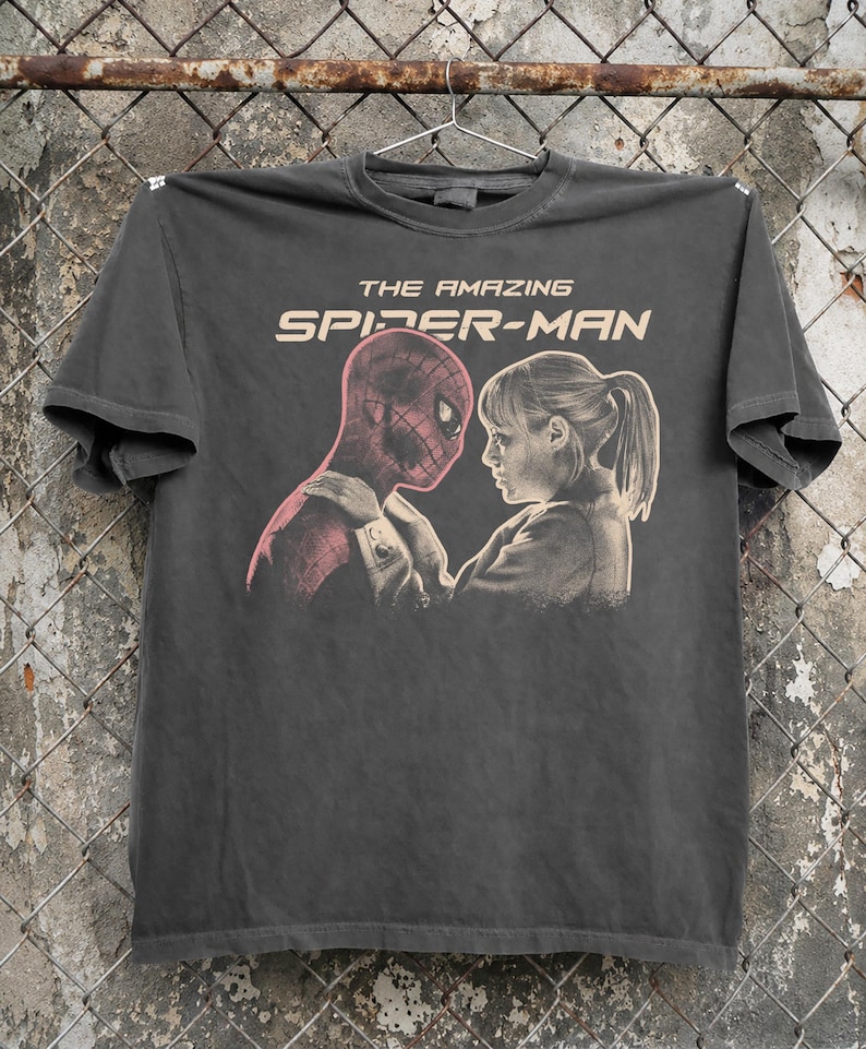 K&ouml;nnte beinhalten: Dunkelgraues T-Shirt mit einer Grafik von Spider-Man und einer Frau, die sich gegen&uuml;berstehen. Der Text "THE AMAZING SPIDER-MAN" ist in hellem Beige &uuml;ber dem Bild aufgedruckt. Das T-Shirt h&auml;ngt an einem Metallb&uuml;gel.