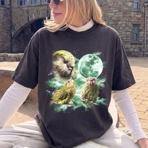 May include: A charcoal grey short-sleeve t-shirt featuring a graphic print of three green kakapo parrots and a large glowing green moon surrounded by green nebula-like clouds. The shirt is worn over a white long-sleeve base layer.