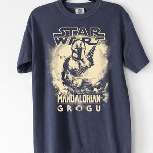 May include: Navy blue t-shirt featuring a graphic from the Star Wars series, with the text "STAR WARS" above an illustration of The Mandalorian holding a weapon, and the text "THE MANDALORIAN AND GROGU" below.