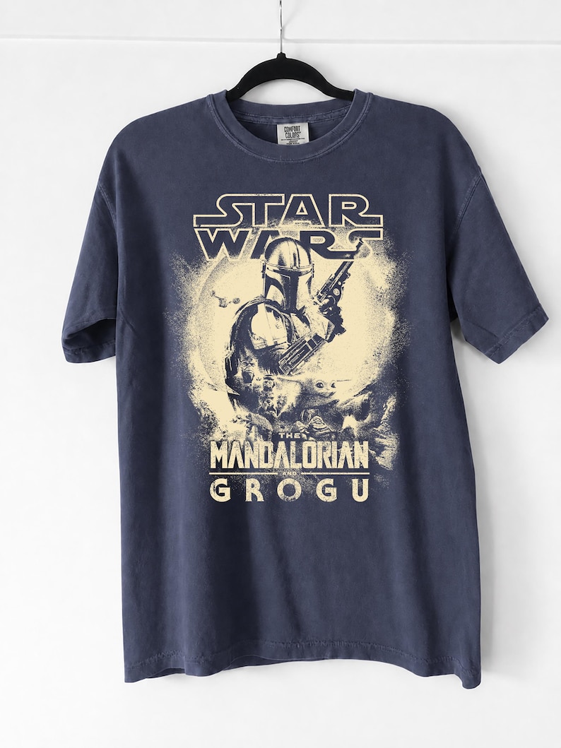 May include: Navy blue t-shirt featuring a graphic from the Star Wars series, with the text "STAR WARS" above an illustration of The Mandalorian holding a weapon, and the text "THE MANDALORIAN AND GROGU" below.