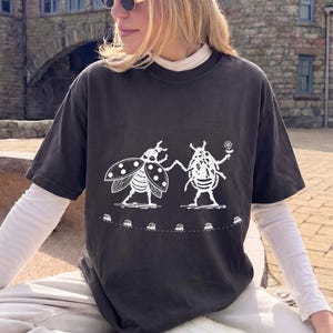 May include: A charcoal gray short-sleeve t-shirt featuring a white graphic print of a ladybug and a bee holding hands, with the bee holding a small flower. Below the insects is a dotted line with small circular shapes, layered over a white long-sleeve shirt.