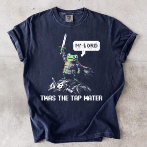 Puede incluir: Una camiseta azul marino oscuro con un gráfico de una rana verde con armadura de colores del arcoíris y una capa, sosteniendo una espada en alto. Un bocadillo pixelado dice M' LORD y el texto debajo de la rana dice TWAS THE TAP WATER.