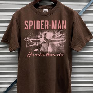 May include: Dark chocolate brown t-shirt featuring the words "SPIDER-MAN" in pink, with an image of Spider-Man and the word "Homecoming" in pink. The shirt is hanging on a wooden hanger.
