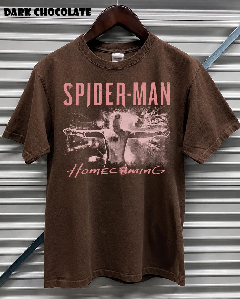 May include: Dark chocolate brown t-shirt featuring the words "SPIDER-MAN" in pink, with an image of Spider-Man and the word "Homecoming" in pink. The shirt is hanging on a wooden hanger.
