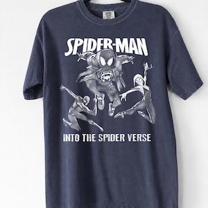 May include: Navy blue t-shirt featuring a graphic of Spider-Man and other Spider-Verse characters. The words "SPIDER-MAN" and "INTO THE SPIDER VERSE" are printed in white. The shirt is hanging on a black hanger.