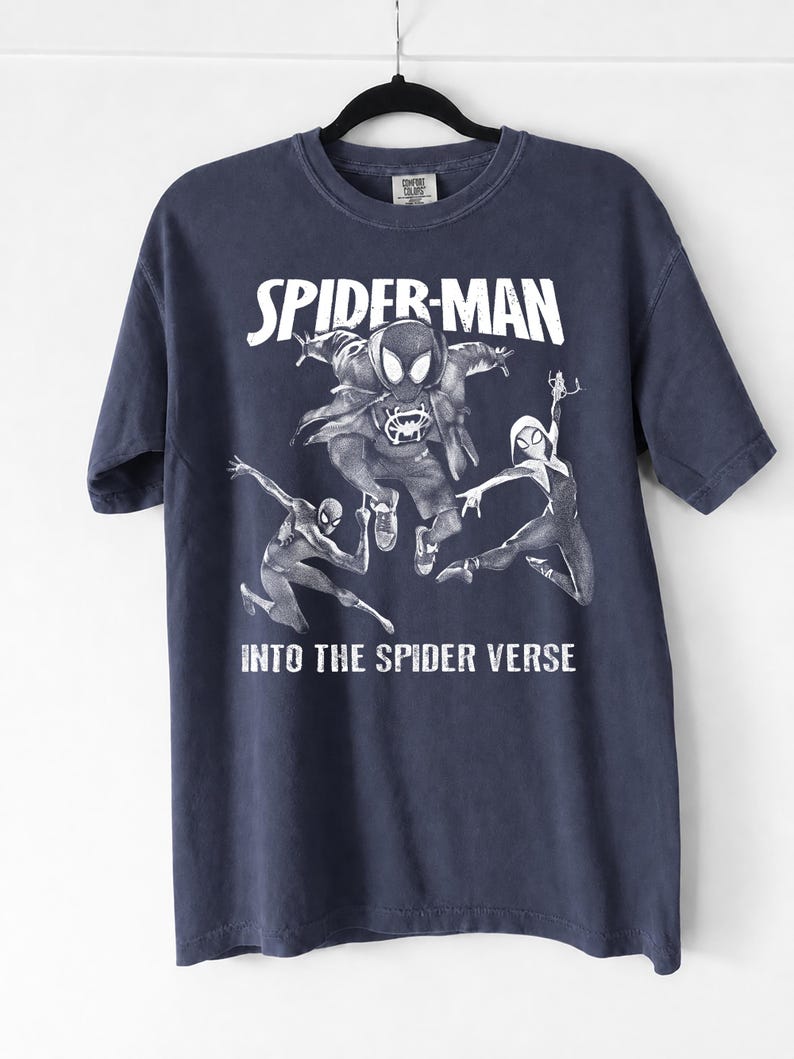 May include: Navy blue t-shirt featuring a graphic of Spider-Man and other Spider-Verse characters. The words "SPIDER-MAN" and "INTO THE SPIDER VERSE" are printed in white. The shirt is hanging on a black hanger.