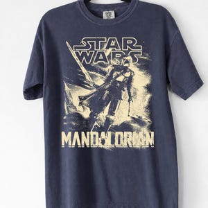 May include: Navy blue t-shirt featuring a distressed graphic of The Mandalorian from Star Wars. The design includes the Star Wars logo and the word "Mandalorian" in a vintage style. The shirt is hanging on a black hanger.